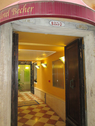Photo shows the entrance under the awning of the Hotel Becher Hotel, with a tiled floor hallway going to a glass door.